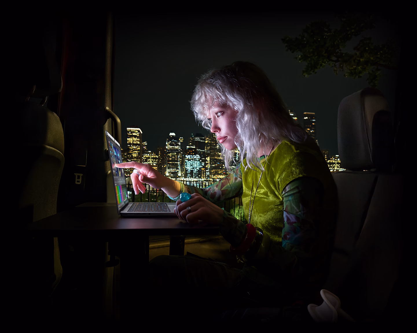 Une personne assise sur la banquette arrière d’une fourgonnette, la porte passager ouverte laisse voir un paysage urbain nocturne, un MacBook Pro avec écran PROLED XDR est posé sur un plan de travail; la personne, éclairée par l’écran, utilise la fonctionnalité tactile pour monter une vidéo dans DaVinci Resolve Studio