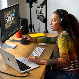 Žena sedí doma u pracovního stolu a používá MacBook Pro připojený k externímu monitoru, má nasazená sluchátka a mluví do mikrofonu