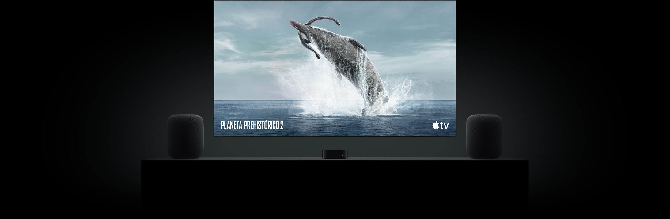 Pantalla de televisión plana y grande en la que se ve una imagen realista de un dinosaurio de «Planeta prehistórico». El televisor está colgado en la pared justo encima de un Apple TV 4K, con dos altavoces HomePod a ambos lados colocados en el mueble del salón
