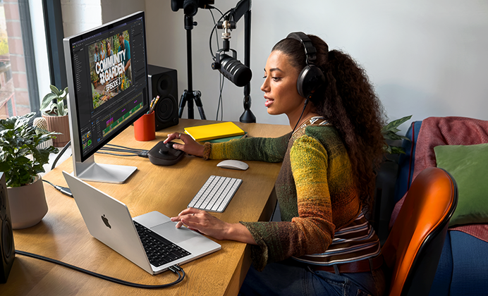 امرأة في منزلها تعمل خلف مكتبها على MacBook&nbsp;Pro متصل بشاشة خارجية وتستخدم سماعات رأس وتتحدث في الميكروفون