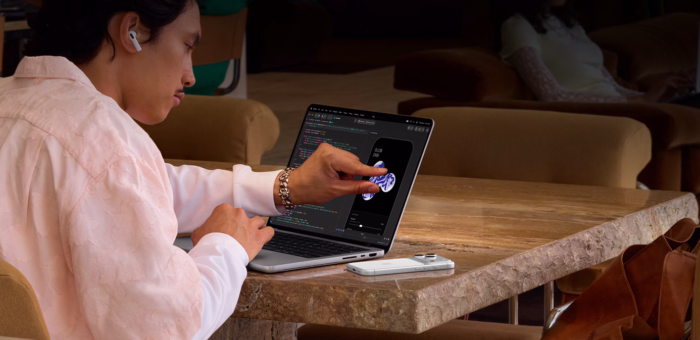 شخص يجلس إلى طاولة في مكان عام، يستخدم إيماءة ضم الإصبعين لتعديل عنصر في تطبيق Xcode باستخدام ميزة اللمس على MacBook Pro مع شاشة PROLED XDR بلون فضي، جهاز iPhone موضوع على الطاولة بجانب اللابتوب والشخص يستخدم سماعات AirPods Pro‏‏