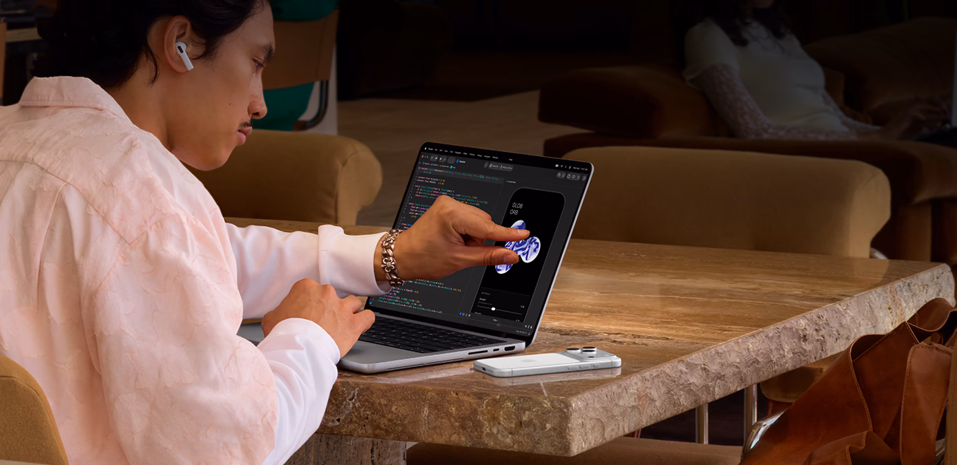 A person sits at a table in a public space, they use a pinch gesture to adjust an element in Xcode using the Touch feature of a silver MacBook Pro with PROLED XDR Display, an iPhone is on the table beside the laptop and the person is using AirPods Pro