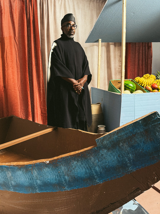 A portrait of A. Potts designer Aaron Potts wearing a long black tunic and a black hat, next to a backdrop made to look like a fruit stand.