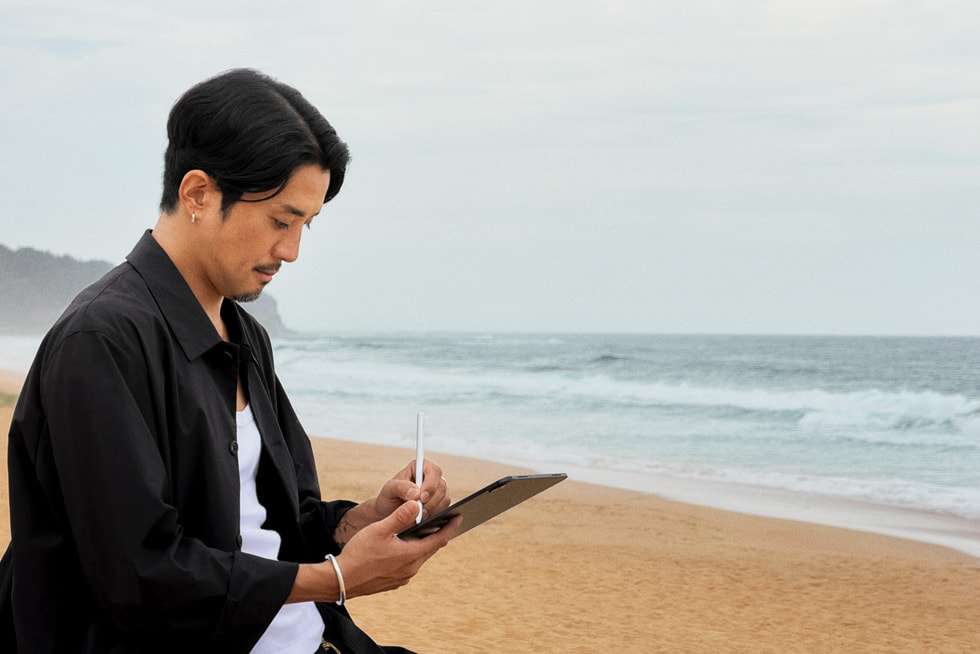 Artist Kentaro Yoshida is shown working with iPad Pro and Apple Pencil while sitting on a beach.