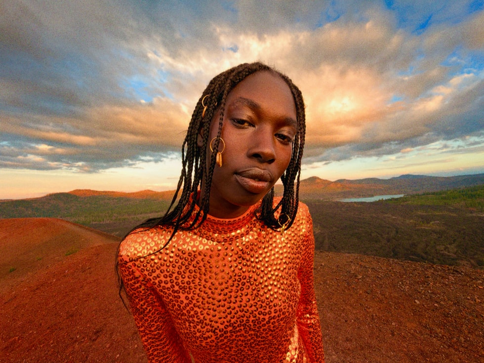 Một người mặc đồ màu cam tạo dáng ở góc chụp cận trung cảnh, phía sau là bầu trời đầy mây và một phong cảnh bao la, rộng lớn.