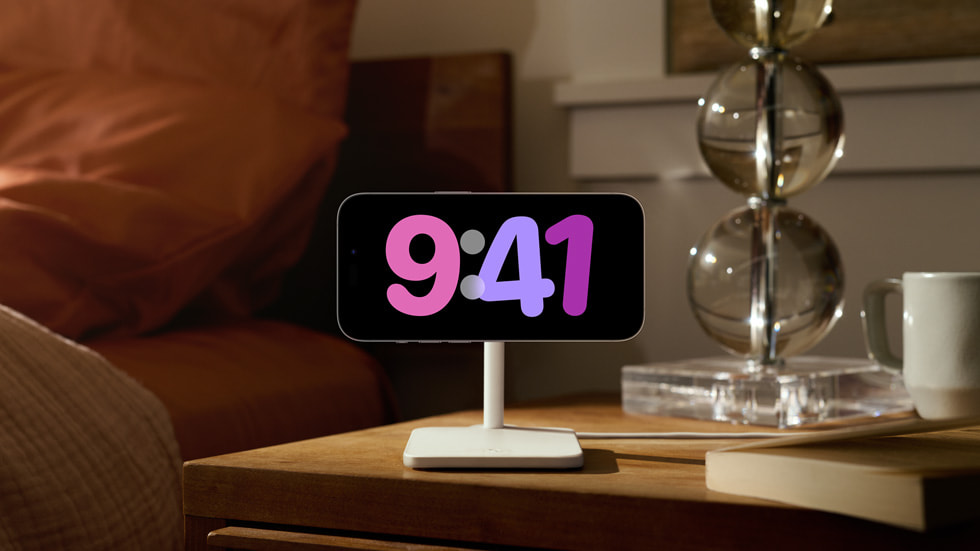 StandBy-oplevelsen med fuld skærm vises på en iPhone 14 Pro, der står på et natbord.