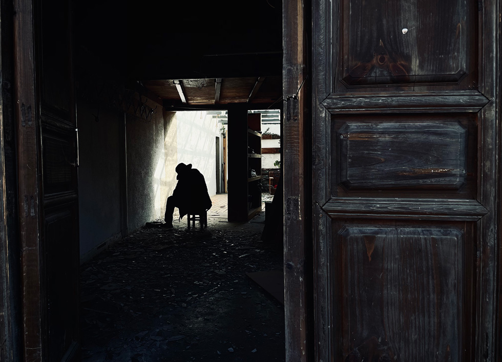 Beyond an opened door, a figure sits amid debris and shadows.