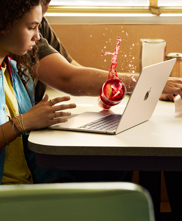 Una persona en un restaurante extiende el brazo para tomar algo de la mesa y tira una bebida sobre una MacBook abierta