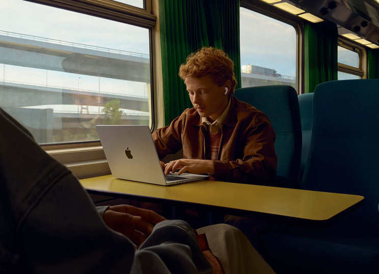 一個人坐在火車靠窗座位上，戴著 AirPods 使用 MacBook 工作