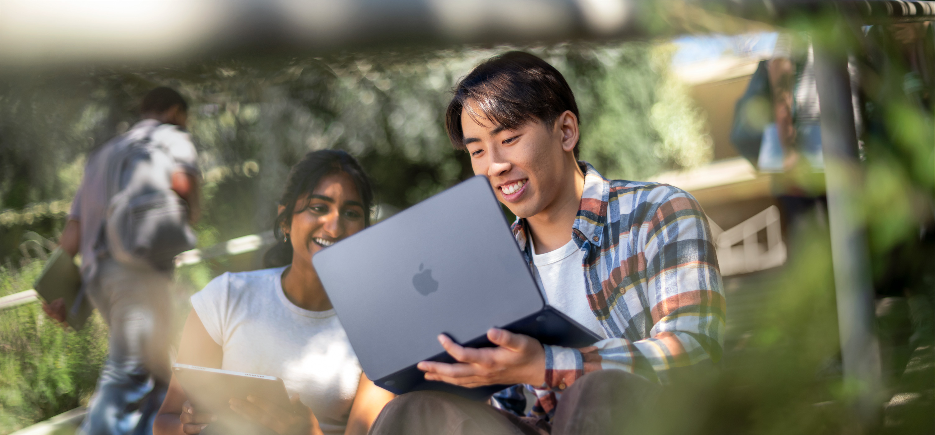 Two smiling students use a MacBook outdoors, one holds it on their lap as they collaborate on an assignment