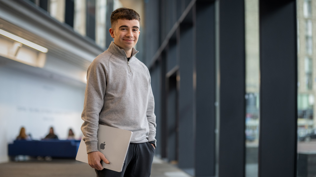 Een student staat in een gang van een modern gebouw met een MacBook-laptop en lacht naar de camera