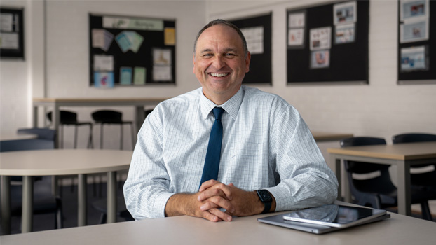 Principal Mark Watkins smiling while sitting at a school desk with a closed MacBook and iPad next to him.