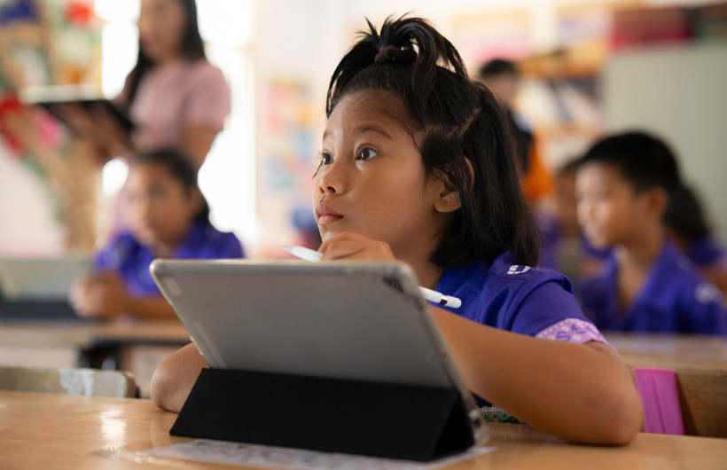 Een jong meisje zit aan haar tafeltje en kijkt geconcentreerd naar voren. Ze heeft een iPad voor haar neus en houdt een Apple Pencil vast.