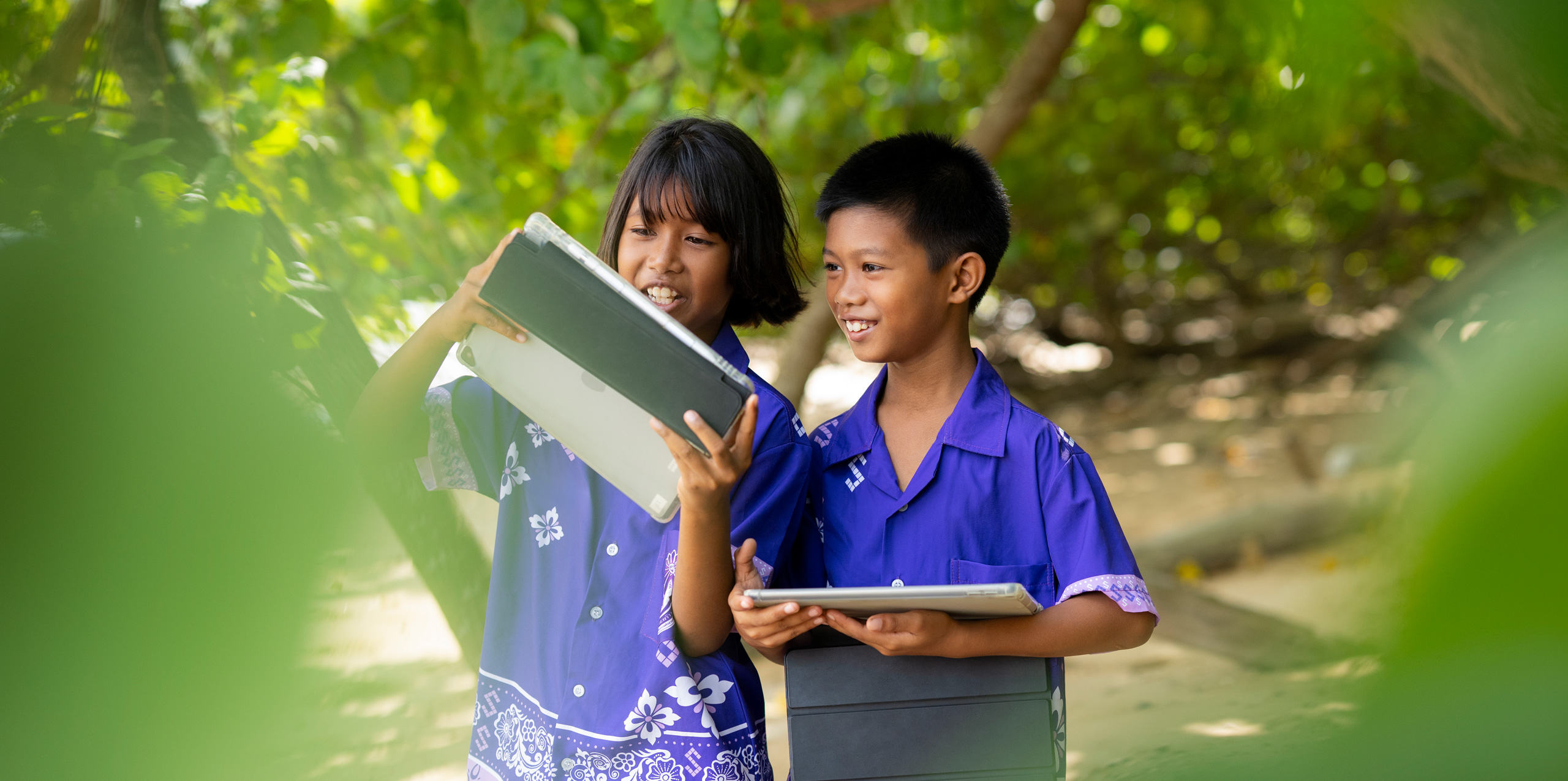 Twee basisschoolleerlingen, een jongen en een meisje, staan buiten tussen tropische bomen en houden allebei een iPad vast. Ze lachen en kijken vol aandacht naar het iPad-scherm van het meisje.