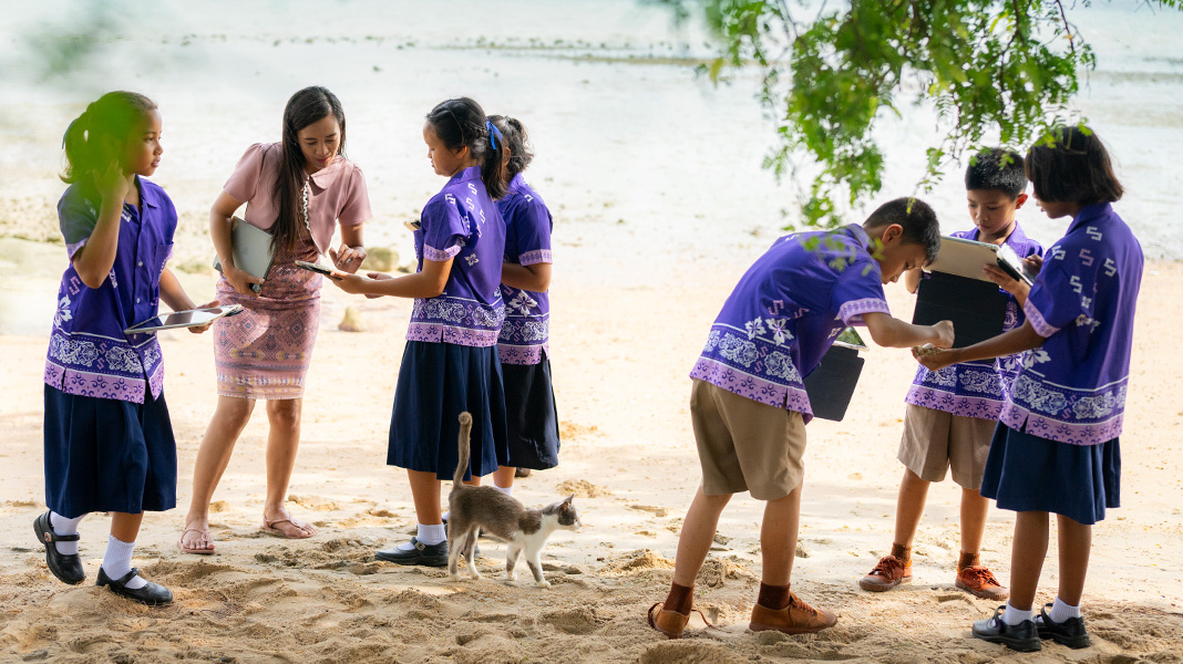 Zes basisschoolleerlingen in een paars uniform zijn op een zandstrand met hun iPad in de weer. De leerkracht helpt een van de leerlingen met haar iPad, en tussen de leerlingen loopt een kat.