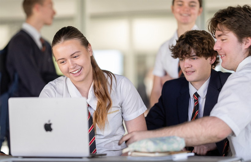 Un groupe d’élèves en uniforme scolaire est réuni autour d’un MacBook, souriant tout en travaillant ensemble.