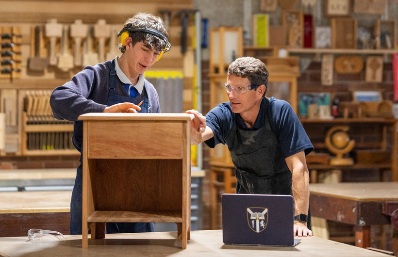 Un enseignant en menuiserie et un élève examinent un petit meuble en bois. Un ordinateur portable avec le logo de l’établissement est posé sur le plan de travail devant eux.
