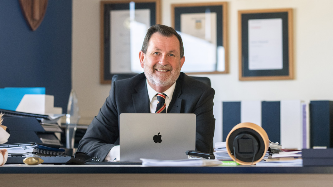Le directeur est assis à son bureau derrière un ordinateur portable Apple ouvert, souriant à la caméra.