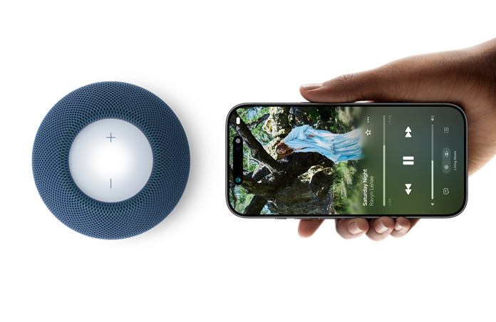 HomePod mini 2 og iPhone: En hånd holder en iPhone ved siden av en blå HomePod mini 2 for Handoff-funksjonen.