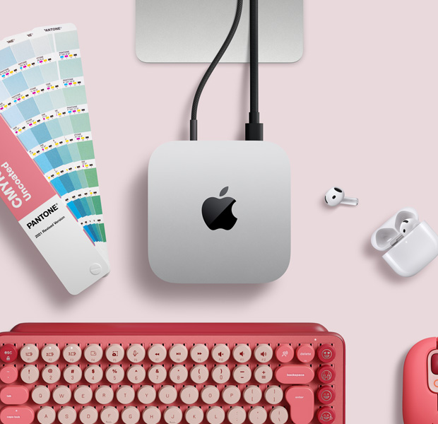 Mac mini, vista superior, muestra el aluminio en plata, las esquinas redondeadas, el logotipo negro y brillante de Apple en el centro, un cable de conexión, parte del soporte del Studio Display, unos AirPods 4, pastillas de colores Pantone y un teclado y ratón Logitech rojos