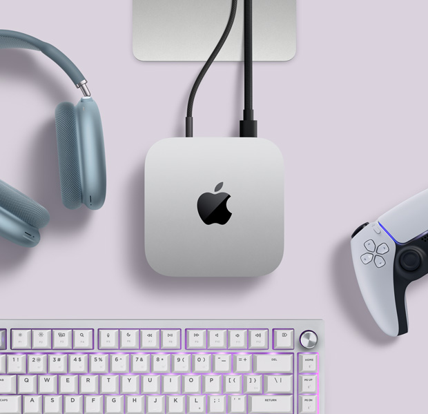 Mac mini, vista superior, muestra el aluminio en plata, las esquinas redondeadas, el logotipo negro brillante de Apple en el centro y un cable de conexión. También se ve parte del soporte del Studio Display y unos AirPods Max 2 en azul, un mando DualSense de PlayStation en blanco y un teclado blanco.