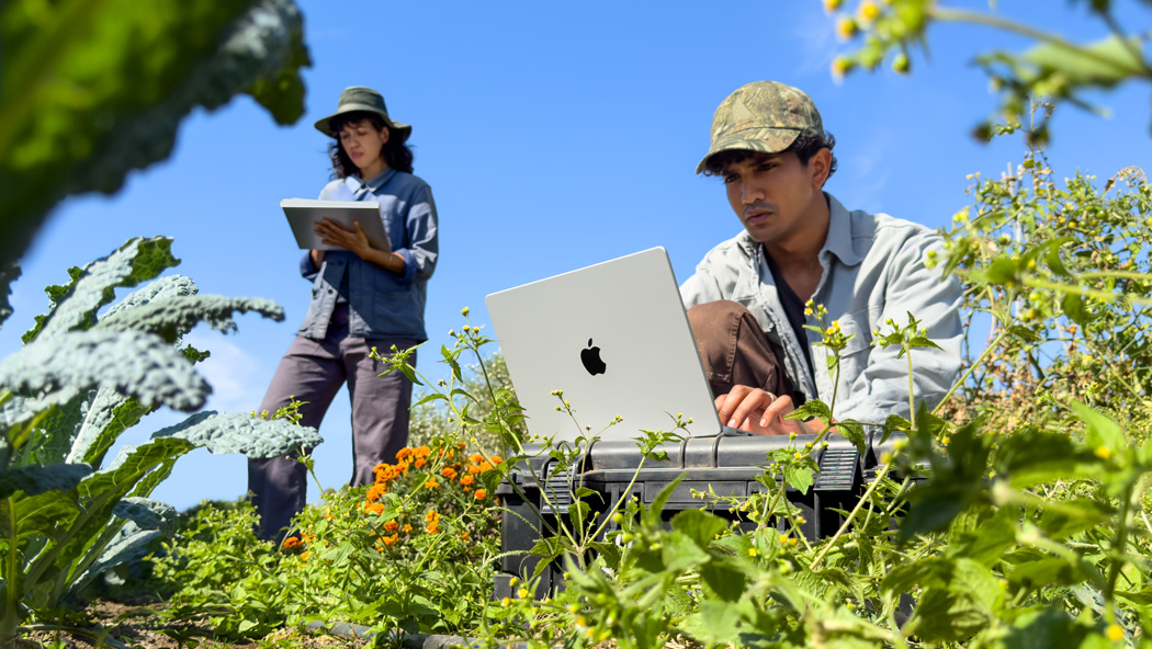兩個人在户外花園工作，一人使用未接上電源的銀色 MacBook Pro，另一人手持筆記簿