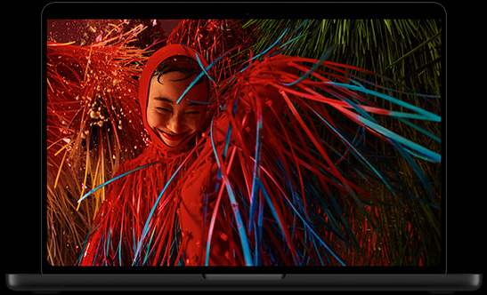 MacBook Pro avec Écran PROLED XDR, noir sidéral, ouvert, l’écran affiche la photo d’une personne souriante vêtue d’une tenue rouge surréaliste avec des détails en plumes
