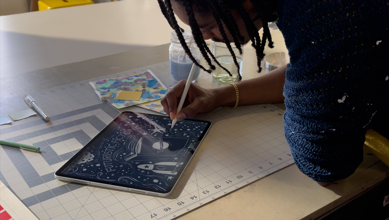 Person with braids wearing a blue sweater drawing on iPad Pro with Apple Pencil, screen shows illustration of a night sky with a planet on a book cover, space ship, butterfly, and futuristic fiction club written in the center