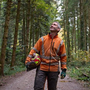 Man wearing an orange high-visibility jacket holding a hard hat while standing on a forest path in a sustainably managed forest in Switzerland
